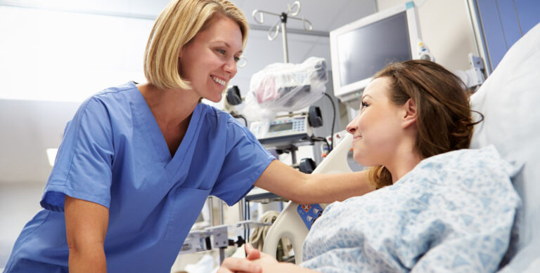 A nurse in blue scrubs is smiling and comforting a patient lying in a hospital bed. The patient, wearing a hospital gown, looks back at the nurse with a slight smile. Medical equipment is visible in the background.