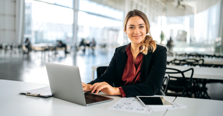 A woman in a red blouse and black blazer sits at a table in a bright, spacious office. She is using a laptop, with a tablet and documents nearby. There are blurred figures in the background. She is smiling at the camera.