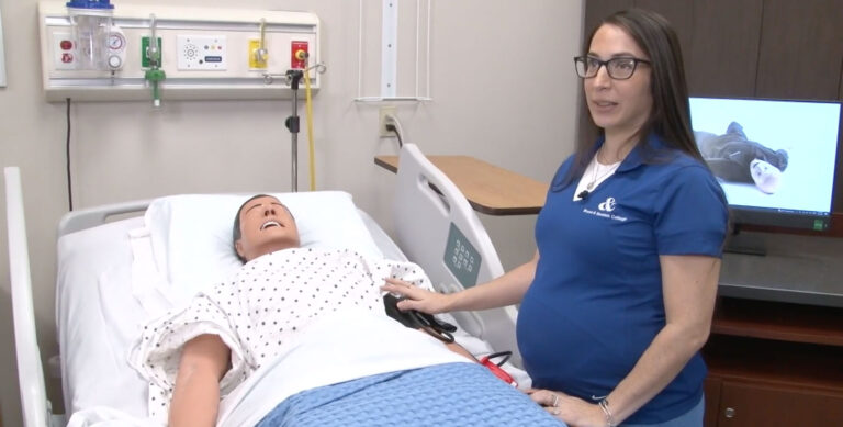 A woman in a blue shirt stands beside a hospital bed, interacting with a medical mannequin. The mannequin is lying on the bed in a gown with a checkered pattern, and a computer screen in the background shows a similar mannequin.